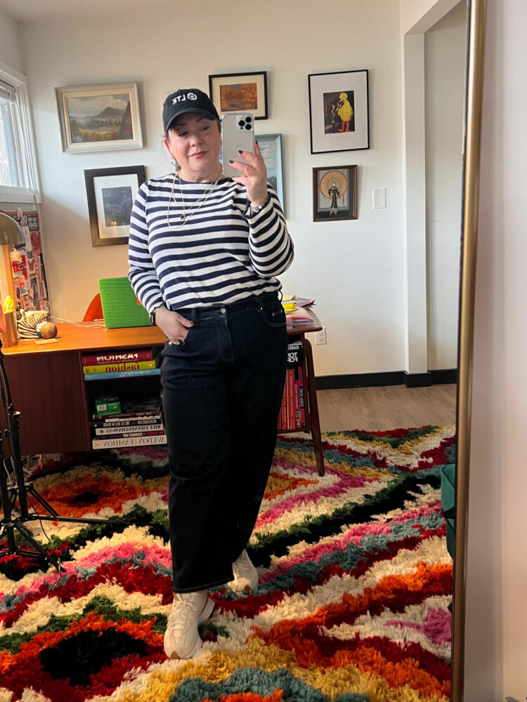 Woman in a black baseball cap, navy and white striped Breton top tucked into the dark selvedge wash of Universal Standard Bae Jeans. New Balance 327 sneakers on her feet.