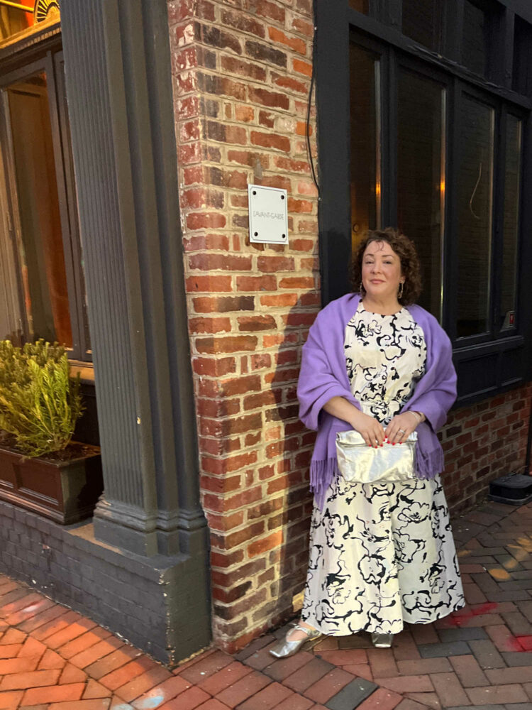 Charles Perri, a petite over-40 woman with curly brown hair is standing outside L'Avant-Garde in Georgetown. She is wearing a black and white patterned puff sleeve top and ankle length skirt. A lavender pashima is over her shoulders and she is holding a silver clutch purse.