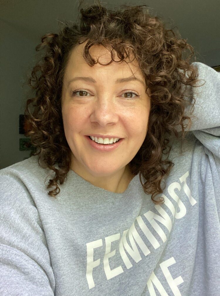 Charles Perri of Wardrobe Oxygen smiling at the camera, not wearing makeup. She is wearing a gray sweatshirt that says Feminist on the front