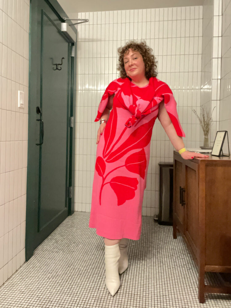 Charles Perri of Wardrobe Oxygen in the Ann Taylor Floral Jacquard sweater tank dress in a pink and red floral. She has a matching cardigan tied around her shoulders and is wearing bone colored pointed toe boots.