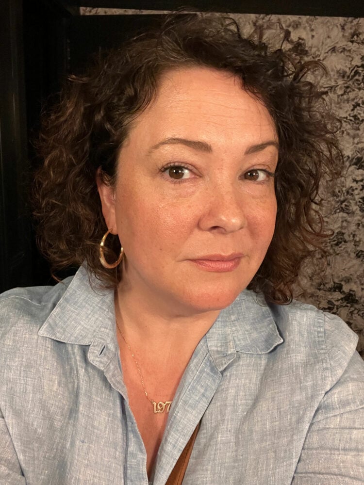 Selfie of Charles Perri wearing a blue linen shirt, gold hoop earrings and a gold necklace with a "1975" charm. She has shoulder length curly brown hair and is looking at the camera