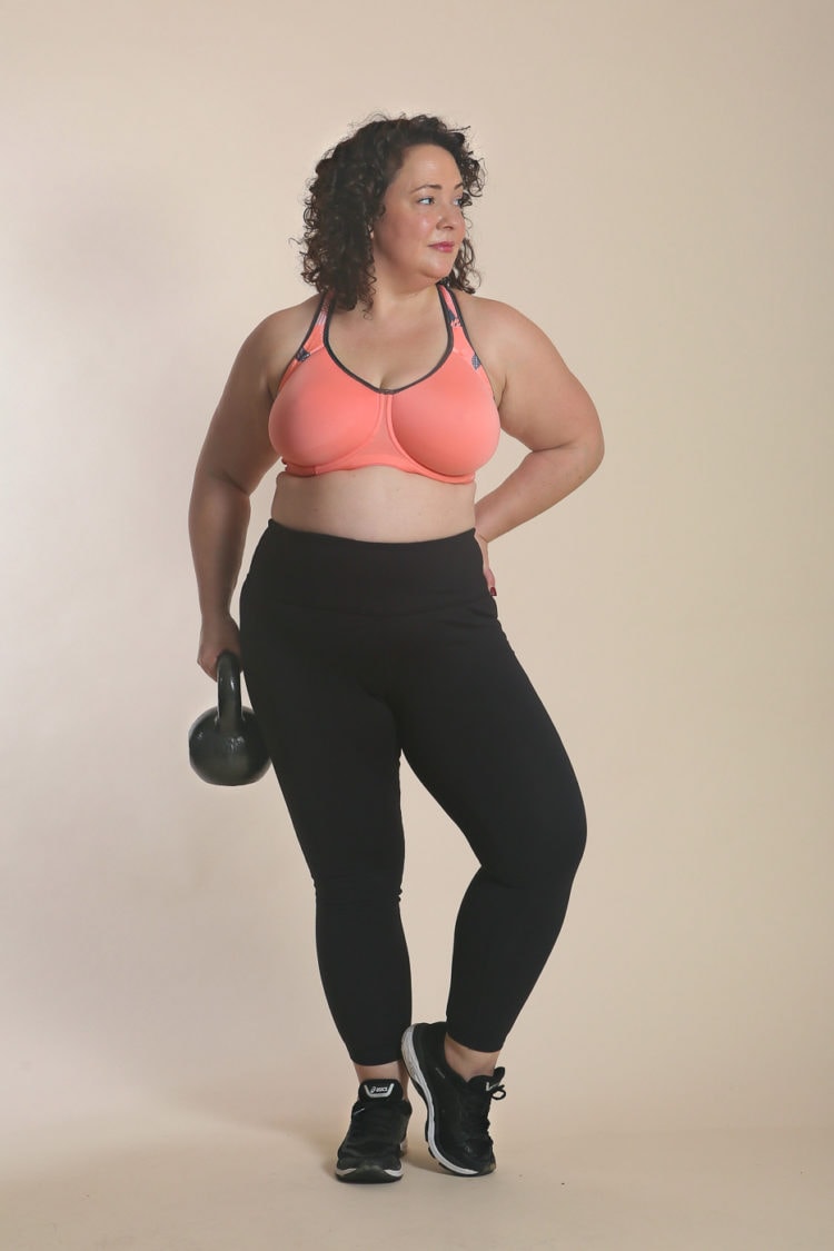 woman in a peach colored sports bra from Wacoal and black leggings. She is holding a kettlebell and waring black Asics sneakers