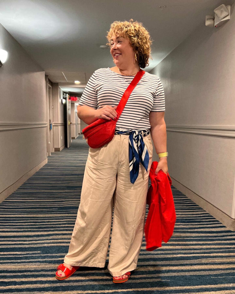 Alison wearing a Breton stripe top tucked into beige Madewell Harlow pants. She has a printed silk scarf tied as a belt. She has a red belt bag worn as a crossbody and is wearing red Birkenstock arizona sandals. A red Chico's Zenergy jacket is in her hand. 