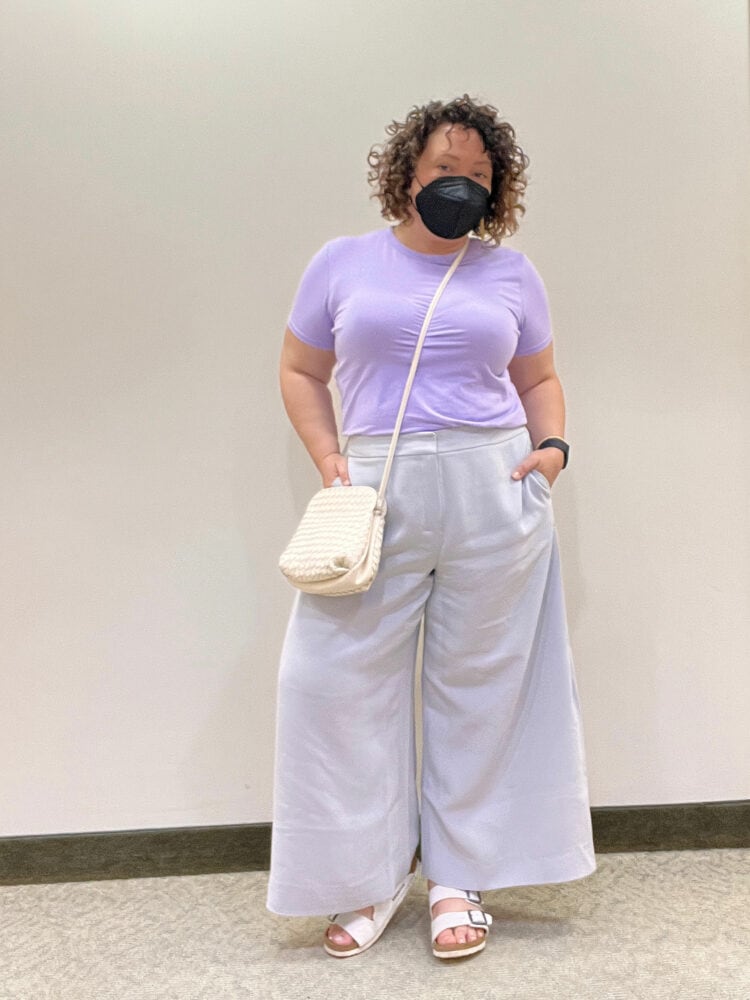 Photo of Charles Perri of Wardrobe Oxygen. She has chin-length curly brown hair and is wearing a lavender short-sleeved t-shirt tucked into pale gray crepe ankle-length wide leg trousers. She has on white Birkenstock sandals and a cream woven leather crossbody bag.