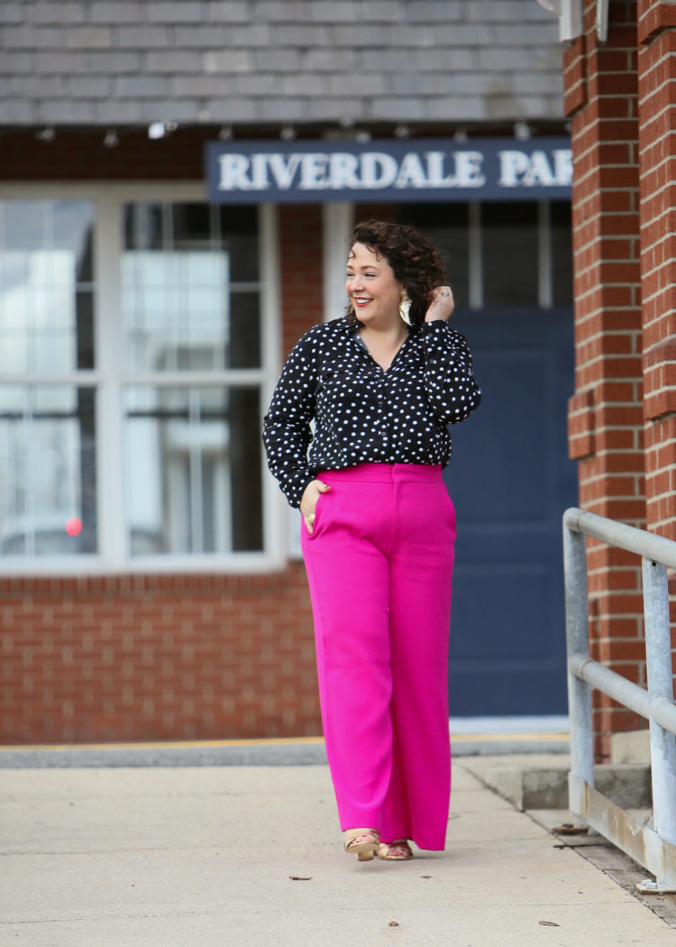 Alison of Wardrobe Oxygen in hot pink wide leg pants and a black blouse with white polka dots tucked into them. She is wearing gold leather heeled sandals from Margaux and is standing in front of a train station in Riverdale, Maryland