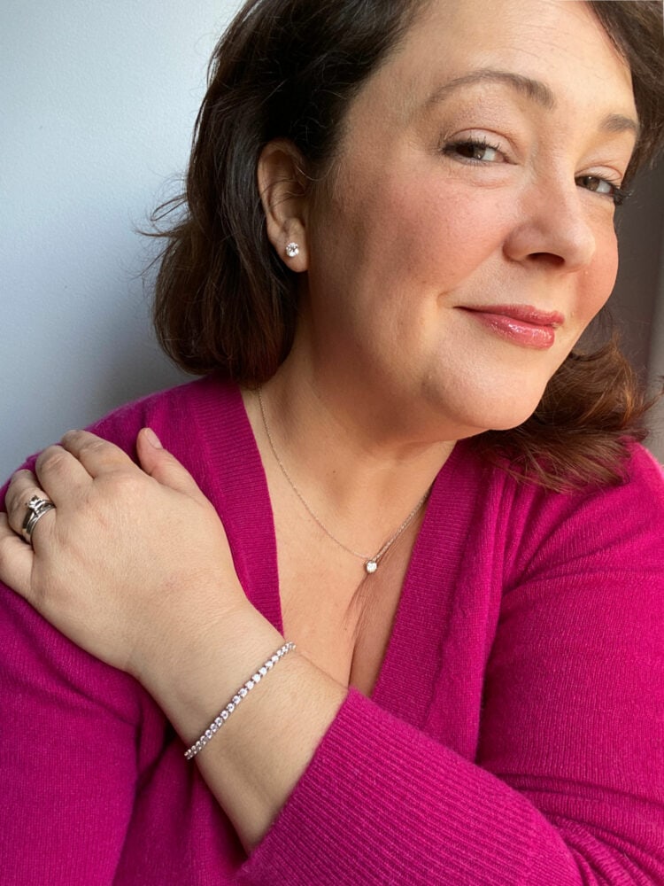 Charles Perri wearing a berry colored cashmere sweater and is looking at the camera with a slight smile. Her left arm is across her body with her hand on her right shoulder.