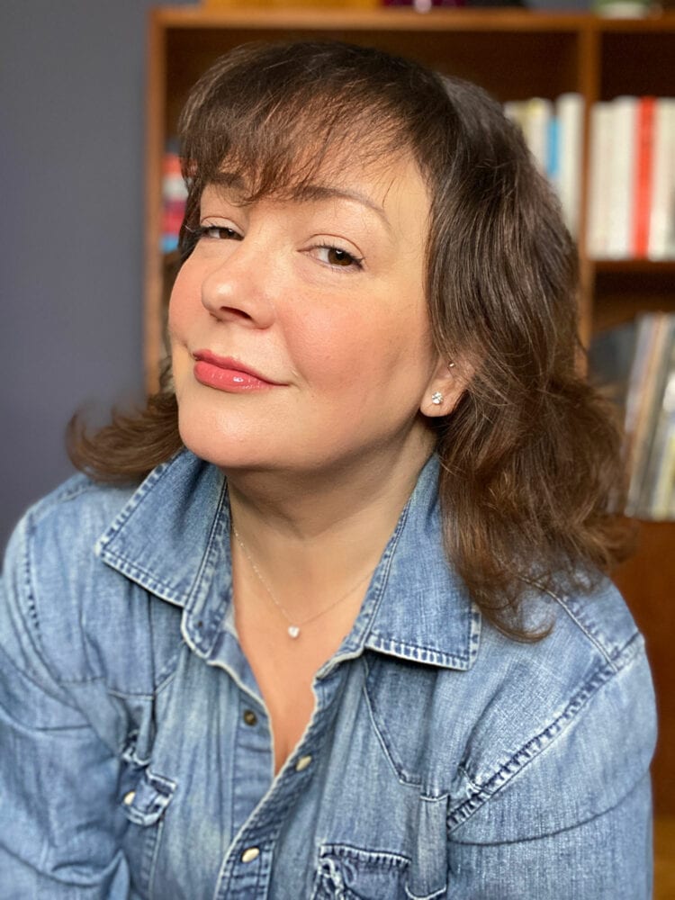 Charles Perri of Wardrobe Oxygen in a denim shirt looking at the camera and smirking. She is wearing James Allen diamond studs in her ears and a delicate silver chain with a diamond pendant on it.