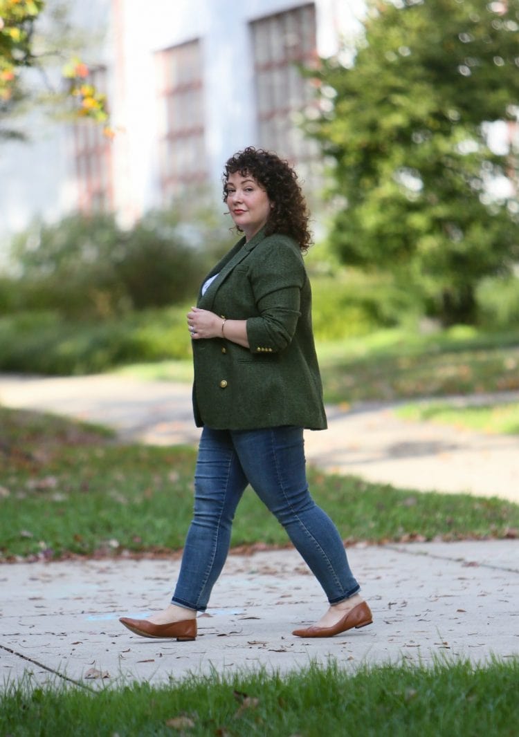 Woman walking down a sidewalk in a green tweed doublebreasted blazer from J. Crew and Universal Standard Seine skinny jeans. She has brown leather flats from Ally NYC on her feet and she turned her head to look at the camera.