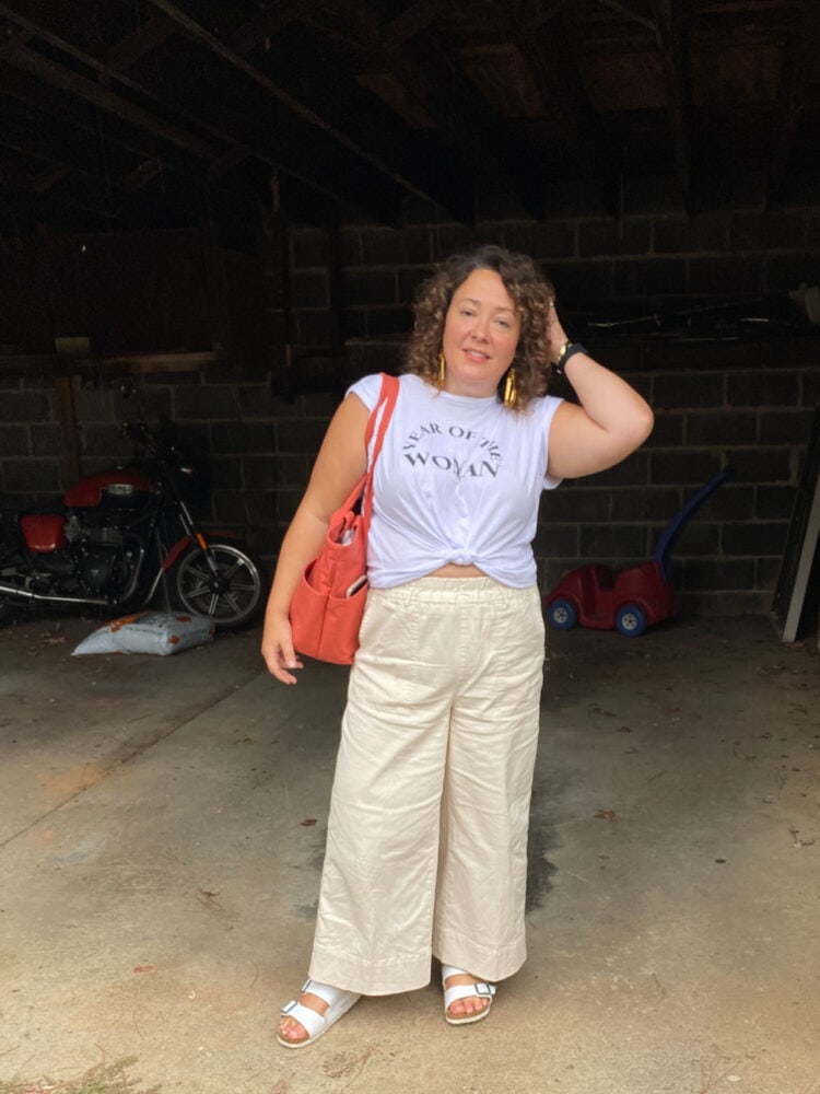 Alison of Wardrobe Oxygen in a white graphic t-shirt knotted over a pair of elastic waist pull-on wide leg chinos from J. Crew. She is wearing Birkenstock sandals and on her shoulder is the Dagne Dover Vida tote in orange organic cotton