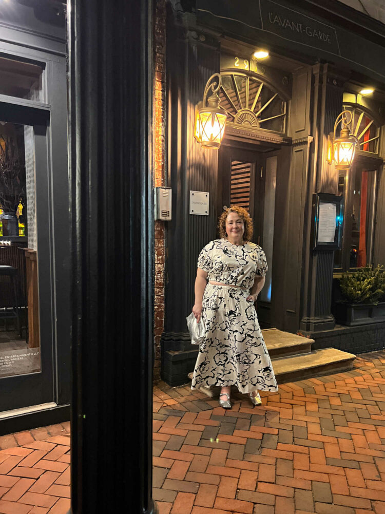 Charles Perri, a petite over-40 woman with curly brown hair is standing outside L'Avant-Garde in Georgetown. She is wearing a black and white patterned puff sleeve top and ankle length skirt. A lavender pashima is over her shoulders and she is holding a silver clutch purse.