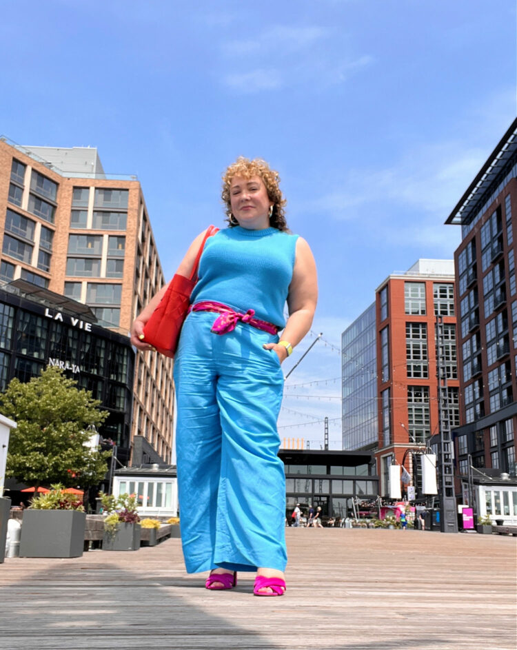 Alison in a blue sleeveless sweateer tucked into the same color blue wide leg linen pants from Ann Taylor. She is standing at the DC Wharf and has an orange leather tote on her shoulder.