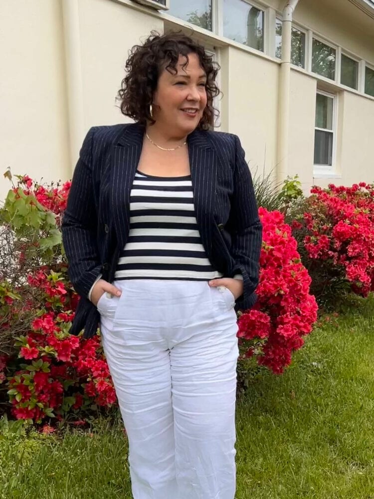 Alison of Wardrobe Oxygen standing outside a cream stucco building with red azalea bushes along the perimter. She is wearing a navy pinstripe blazer, navy and white stripe scoop neck tank, and white linen pants. Her hands are in her pants pockets and she is looking away from the camera.