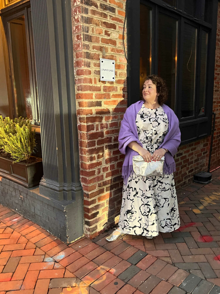 Charles Perri, a petite over-40 woman with curly brown hair is standing outside L'Avant-Garde in Georgetown. She is wearing a black and white patterned puff sleeve top and ankle length skirt. A lavender pashima is over her shoulders and she is holding a silver clutch purse.