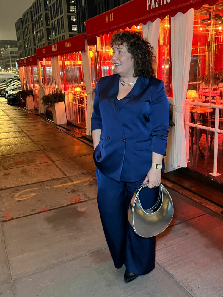 Charles Perri, a petite size 14/16 woman over 40, is standing outside of Pastis DC in front of the outdoor dining area. She is in a navy satin double breasted pantsuit from Ann Taylor and is holding a shiny STAUD Moon bag in her hand. It is night and the sidewalk is damp from a recent rain.