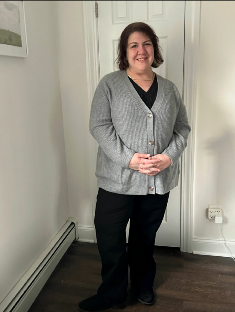 Woman in black pants and the Quince Mongolian Cashmere Oversized Boyfriend Cardigan in Extended Sizes in gray. She has her hands clasped in front of her and is smirking at the camera, standing in a hallway.