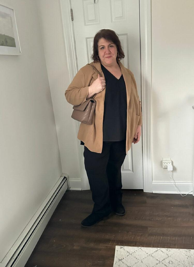 woman wearing a camel sweater blazer from Quince over a black v-neck and black pants. She has a taupe quilted handbag on her shoulder and she looks at the camera while holding the strap of the handbag.