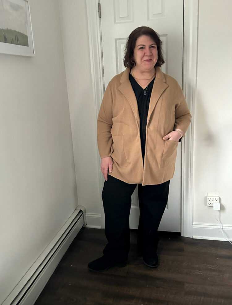 woman with shoulder length brown hair, wearing black pants, a black v-neck top, and a camel sweater blazer from Quince. She has one hand in her pocket and is smiling at the camera.