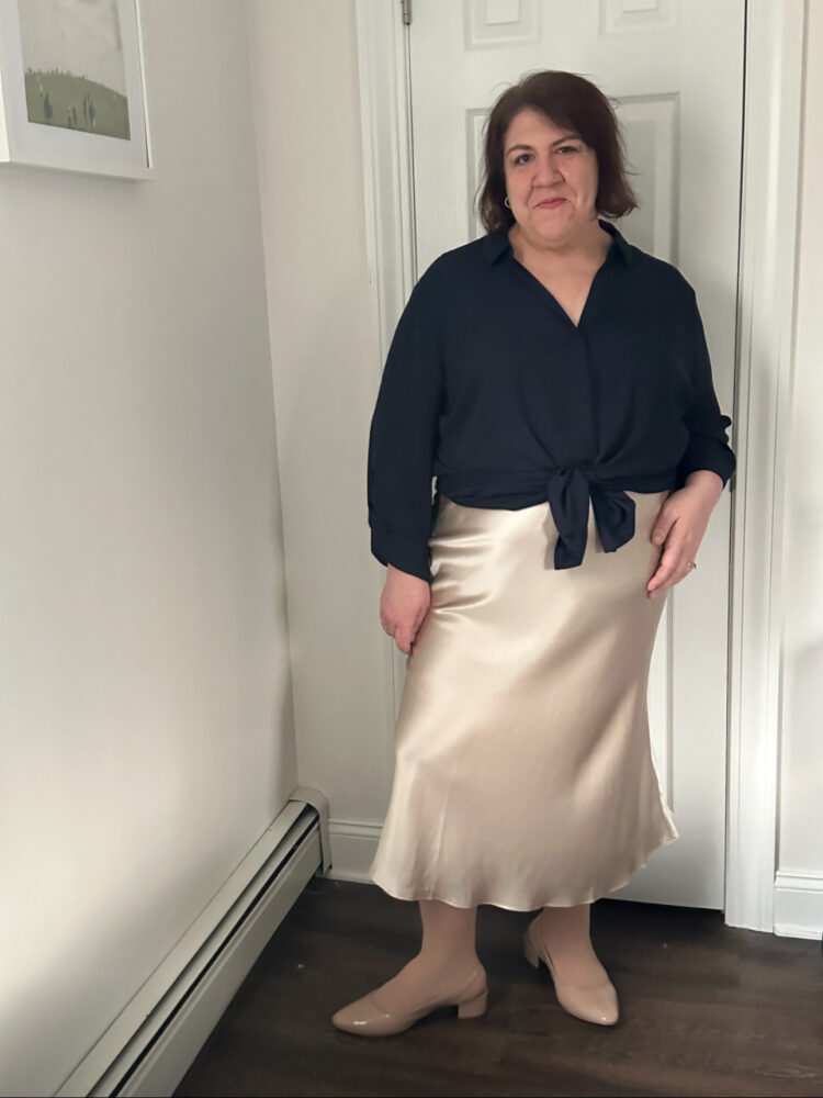 Woman in the Quince plus size washable silk skirt in champagne with a black blouse tied at the waist. She is standing in a hallway and smiling at the camera.