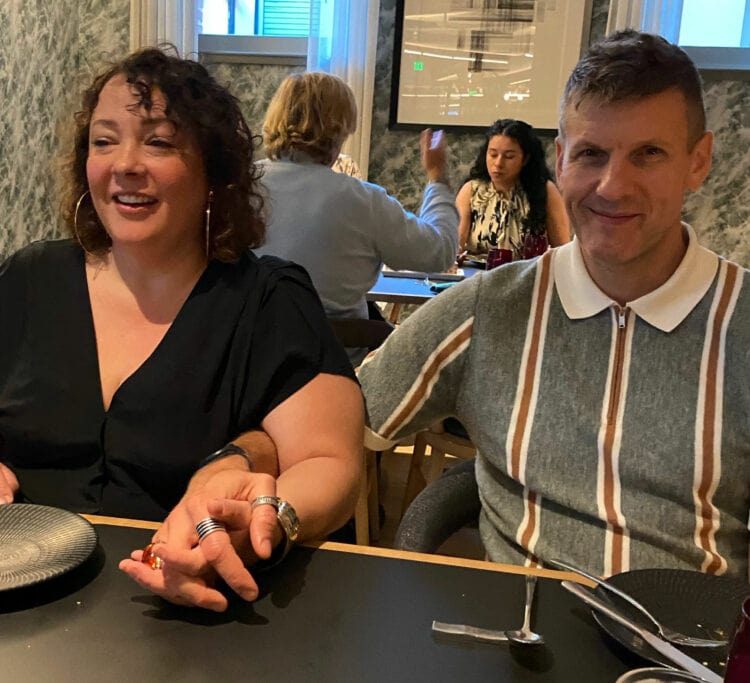 Alison and Karl Gary sitting next to each other at a table in a restaurant, holding hands. Alison has limp curly hair that falls to her shoulders.