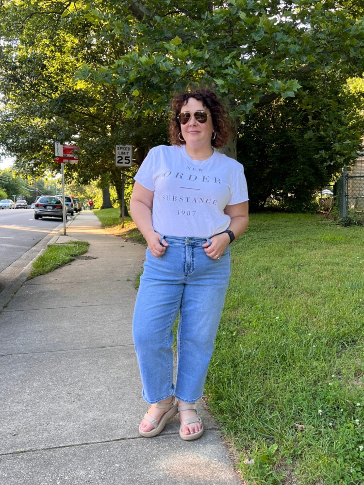 Woman in a New Order t-shirt and a faded pair of the Universal Standard Bae cropped jeans. She is wearing white Birkenstock sandals and walking down a sidewalk in a suburban neighborhood.