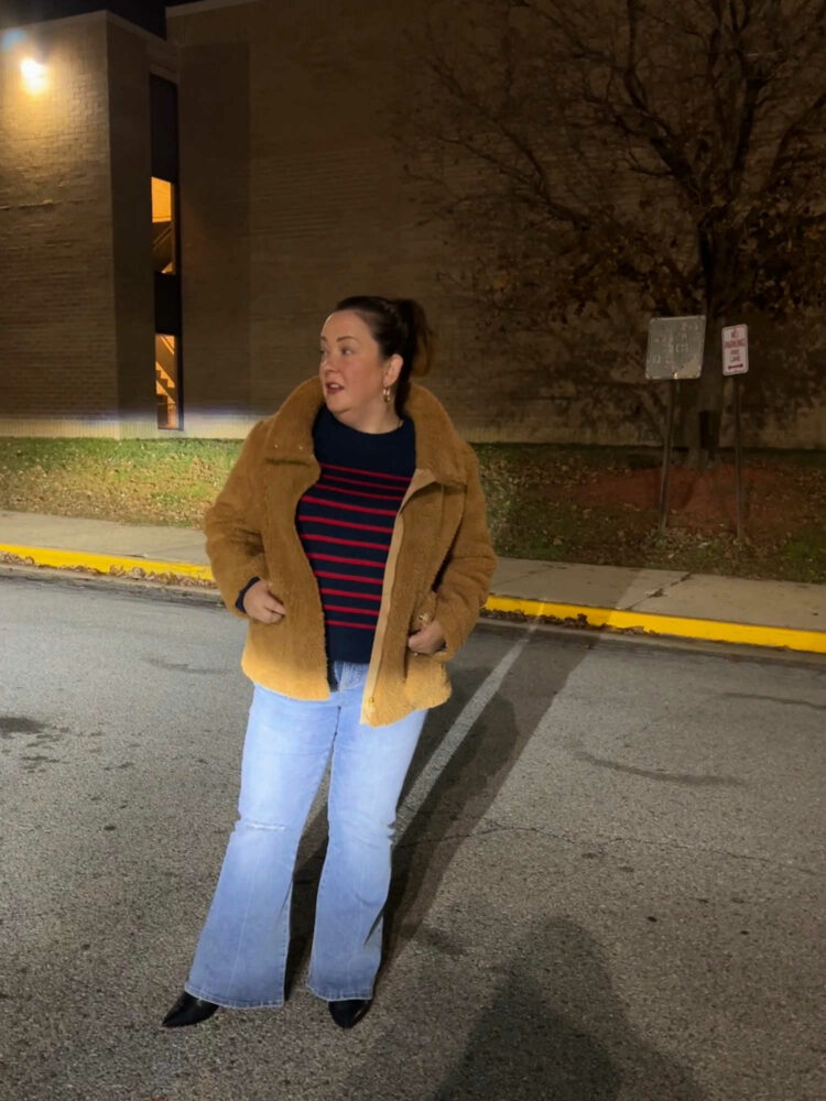 woman standing in a parking lot at night, the headlights of a car illuminating her. She is wearing a tan teddy short coat, a navy sweater with red stripes, and a faded pair of the Universal Standard Farrah flared jeans