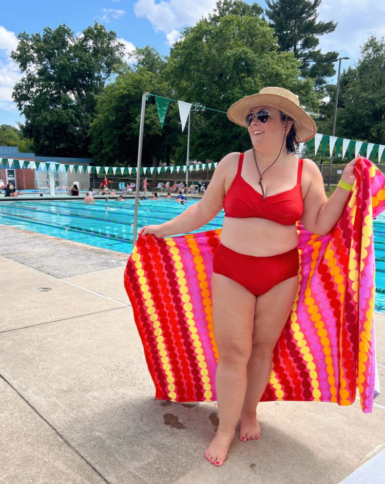 Woman in a red large bust bikini holding a printed towel standing at a swimming pool