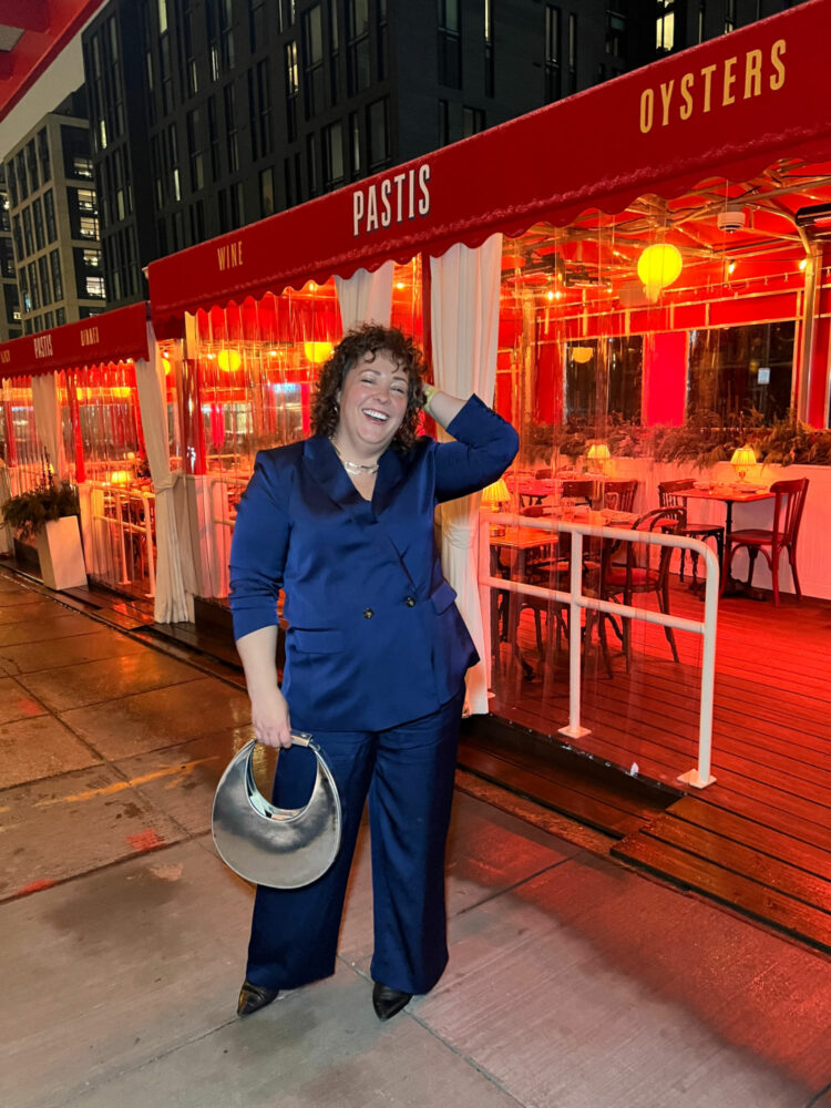 Charles Perri of Wardrobe Oxygen standing in front of the restaurant Pastis in the Union Market district of Washington DC. She is wearing a navy satin pantsuit. One hand is in her hair, the other is holding a silver chrome STAUD moon bag. She is laughing.