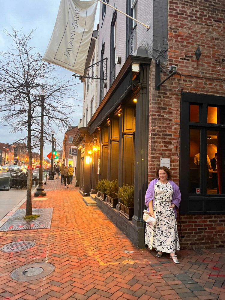 Charles Perri, a petite over-40 woman with curly brown hair is standing outside L'Avant-Garde in Georgetown. She is wearing a black and white patterned puff sleeve top and ankle length skirt. A lavender pashima is over her shoulders and she is holding a silver clutch purse.