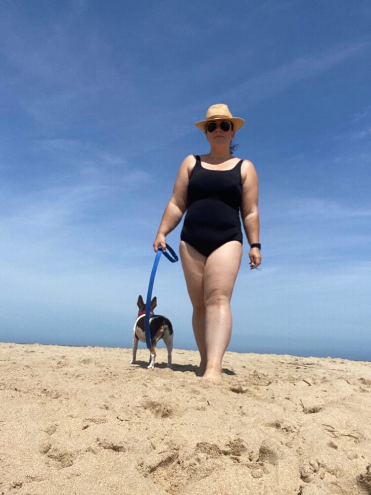 Wardrobe Oxygen wearing the Lands' End Tugless Tank in black with a straw fedora and aviator sunglasses. She is standing on the beach with her dog on a leash.